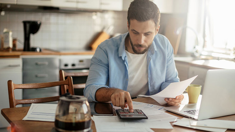 A man reviewing his finances at the table surrounded by papers and a laptop