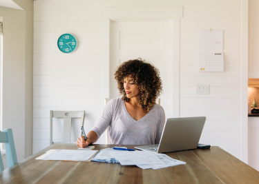 Woman sitting at kitchen table with laptop and several documents spread out